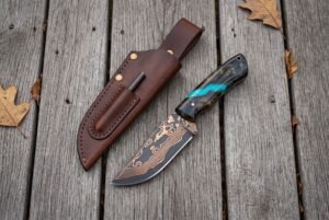 Copper Damascus skinner knife with turquoise accent resin handle lying next to a brown leather sheath on grey planks