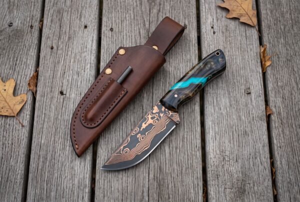 Copper Damascus skinner knife with turquoise accent resin handle lying next to a brown leather sheath on grey planks