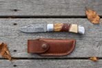 Damascus folding knife with olive wood, red wood and bone handle shown open above its snap closure leather pouch on grey boards