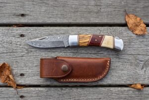 Damascus folding knife with olive wood, red wood and bone handle shown open above its snap closure leather pouch on grey boards