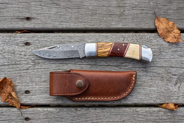 Damascus folding knife with olive wood, red wood and bone handle shown open above its snap closure leather pouch on grey boards