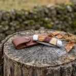 Damascus folding pocket knife with olive wood, red wood and bone handle beside its tan leather pouch on a tree stump