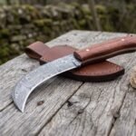 Damascus steel curved blade knife with rosewood handle on its brown leather sheath, laid on old wood beside a jute rope
