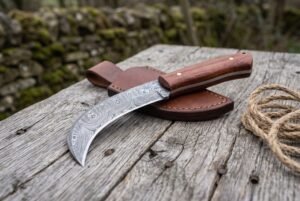 Damascus steel curved blade knife with rosewood handle on its brown leather sheath, laid on old wood beside a jute rope