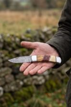Damascus steel folding knife with three part wood and bone handle and steel bolsters held open on a palm outdoors