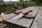 Skinner knife with wood handle resting on its tan leather sheath on an outdoor picnic table beside a dry stone wall