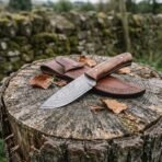 Skinner knife with wood handle on a tree stump with wood shavings, brown leather sheath behind it and a stone wall beyond