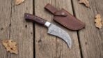 Damascus hawkbill knife with pakka wood handle laid beside its brown leather belt sheath on weathered boards with oak leaves
