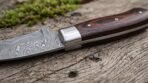 Close up of Damascus hawkbill blade pattern meeting a polished steel bolster and red brown pakka wood handle on rough wood
