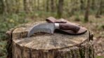 Damascus hawkbill knife with pakka wood handle standing on a tree stump in front of its brown leather sheath in woodland