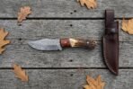 Damascus steel skinner knife with rosewood and stag handle on grey boards from above, stitched leather sheath to the right