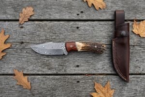 Damascus steel skinner knife with rosewood and stag handle on grey boards from above, stitched leather sheath to the right