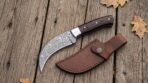 Hooked Damascus hawkbill blade with steel bolster and dark pakka wood scales next to a stitched leather sheath on old planks