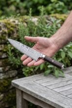 Damascus chef knife with dark wenge wood handle held flat on an open hand above a garden table