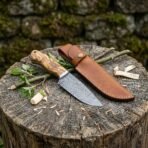 Damascus steel camp knife with olive wood handle on a tree stump beside its tan leather sheath and green twigs