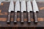 Five Damascus kitchen knives with dark wood handles lined up side by side on a striped end grain board