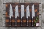 Five Damascus kitchen knives in a row on a wooden board from above, with rosemary and a red onion beside them