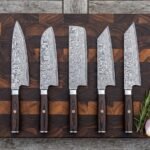 Five Damascus kitchen knives in a row on a wooden board from above, with rosemary and a red onion beside them
