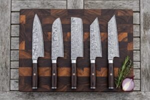 Five Damascus kitchen knives in a row on a wooden board from above, with rosemary and a red onion beside them