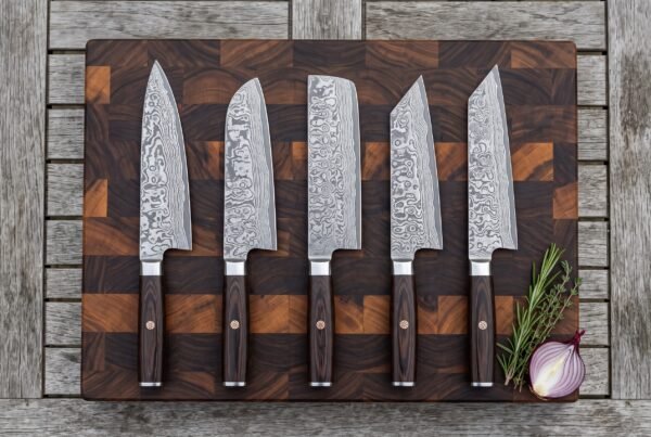 Five Damascus kitchen knives in a row on a wooden board from above, with rosemary and a red onion beside them