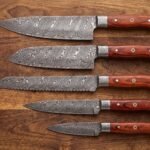 Five Damascus kitchen knives with rosewood handles and mosaic pins laid in a row on a dark wood table