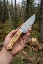 Olive wood handled Damascus camp knife held in one hand in a pine forest with a stack of firewood behind