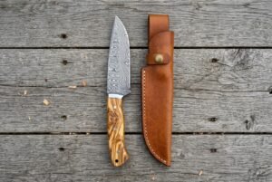 Overhead view of the Damascus camp knife with olive wood handle standing beside its tan leather sheath on boards