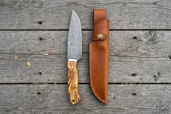 Overhead view of the Damascus camp knife with olive wood handle standing beside its tan leather sheath on boards