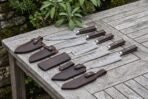 Row of Damascus chef knives with dark wood handles laid out on a garden table, each next to a leather sheath