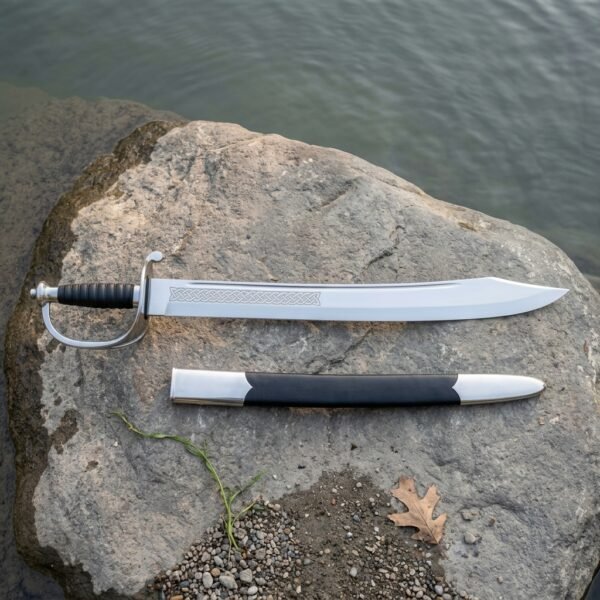 Hand forged naval cutlass with a broad polished steel blade, etched knotwork band, steel stirrup knuckle bow and ribbed black leather grip lying on a river smoothed granite rock beside its black scabbard