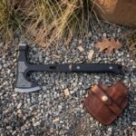 Grimhawk hand forged tactical tomahawk in blackened 1095 carbon steel with hammer poll and skeletonised handle lying on gravel beside its leather belt sheath