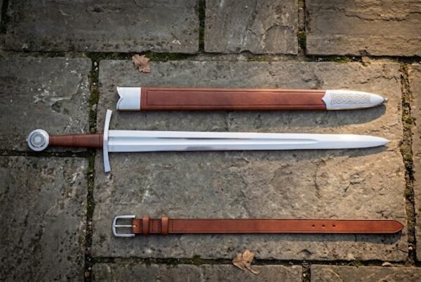 Hand forged arming sword with scabbard and belt, polished fullered type XII blade and steel disc pommel, laid out on mossy grey flagstone paving.