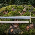 Hand forged elven longsword with an engraved double edged blade and cream leather scabbard lying across a moss covered granite boulder in daylight