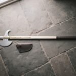Hand forged halberd with a polished steel spike, crescent blade and rear fluke on an ash haft with brass langets lying on grey flagstones by a leather cover