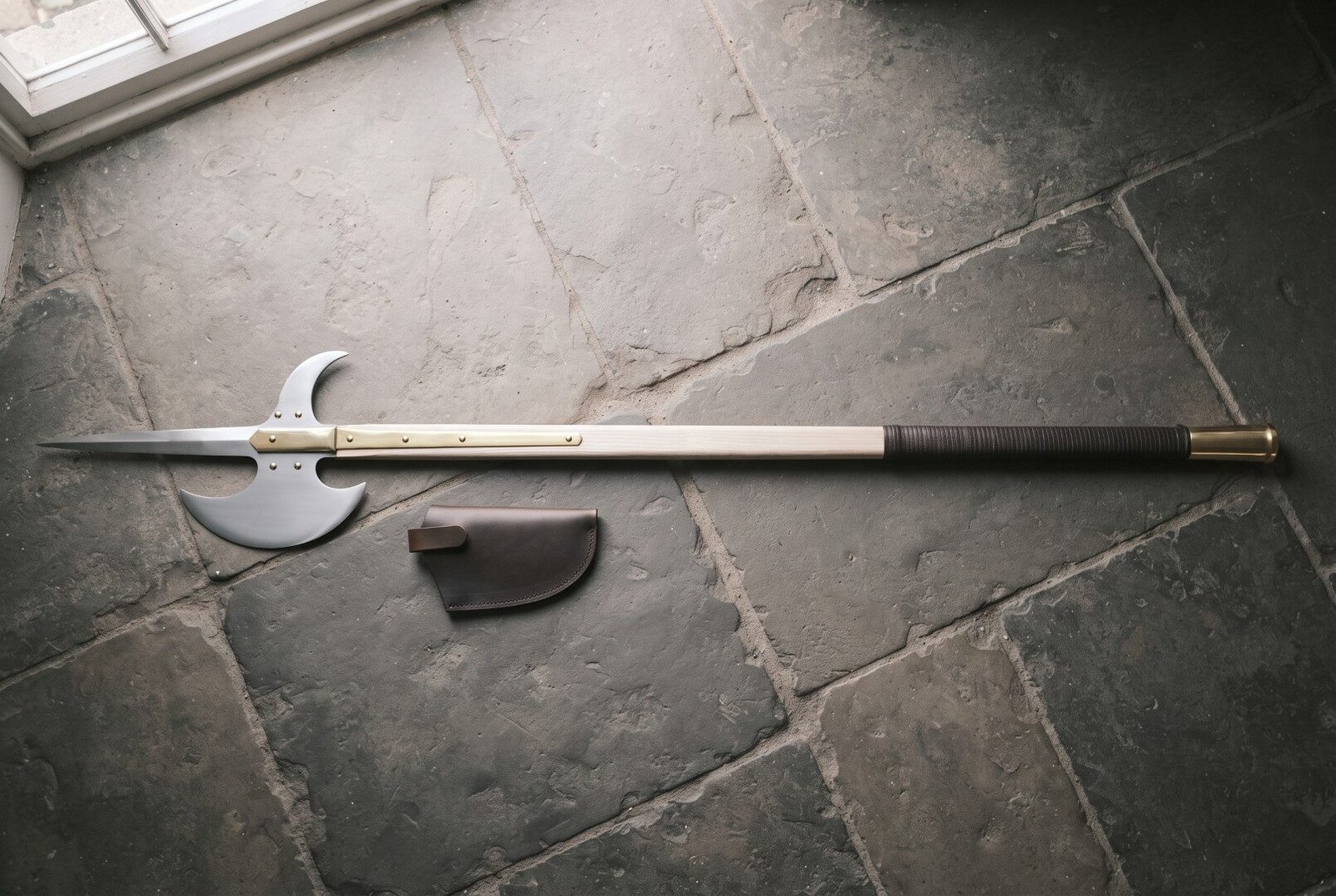 Gemini_Generated_Image_2wqbin2wqbin2wqb Hand forged halberd with a polished steel spike, crescent blade and rear fluke on an ash haft with brass langets lying on grey flagstones by a leather cover