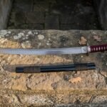 Hand forged short samurai sword with a deep red cord wrapped grip on a weathered stone step, its black saya with brown cord wrap laid below the polished blade