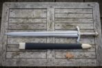 Hand forged elven sword replica and its black leather scabbard laid on weathered grey timber decking, showing the engraved fuller and ribbed grip