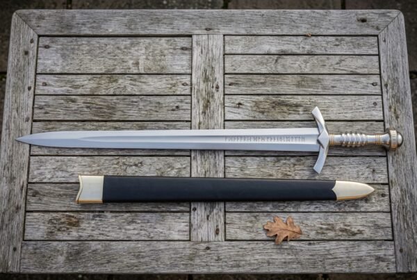 Hand forged elven sword replica and its black leather scabbard laid on weathered grey timber decking, showing the engraved fuller and ribbed grip