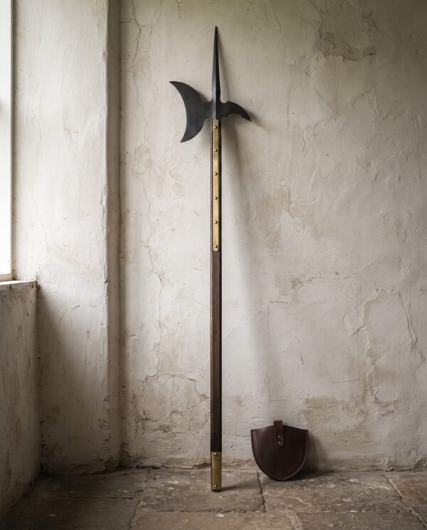 Blackened halberd polearm with an axial spike and crescent blade on a walnut haft with brass langets standing against a pale cracked plaster wall