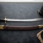 Hand forged curved sword with a broad polished scimitar blade, brass cross guard and leather grip, laid on dark slate beside its tooled leather scabbard and belts