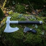 Hand forged norse runic axe with a broad crescent bit, frost blue Elder Futhark runes and a dark leather wrapped haft, resting on a mossy woodland boulder.