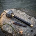 Damascus morning star mace with a forge welded spiked head, brass collars and dark hardwood haft resting on a river smoothed rock at the water edge