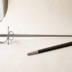 Hand forged rapier with a polished side ring guard and wire wrapped grip photographed on a pale studio backdrop beside its black leather scabbard