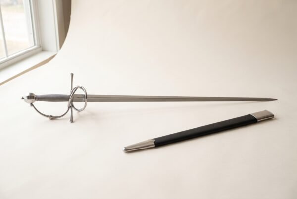 Hand forged rapier with a polished side ring guard and wire wrapped grip photographed on a pale studio backdrop beside its black leather scabbard