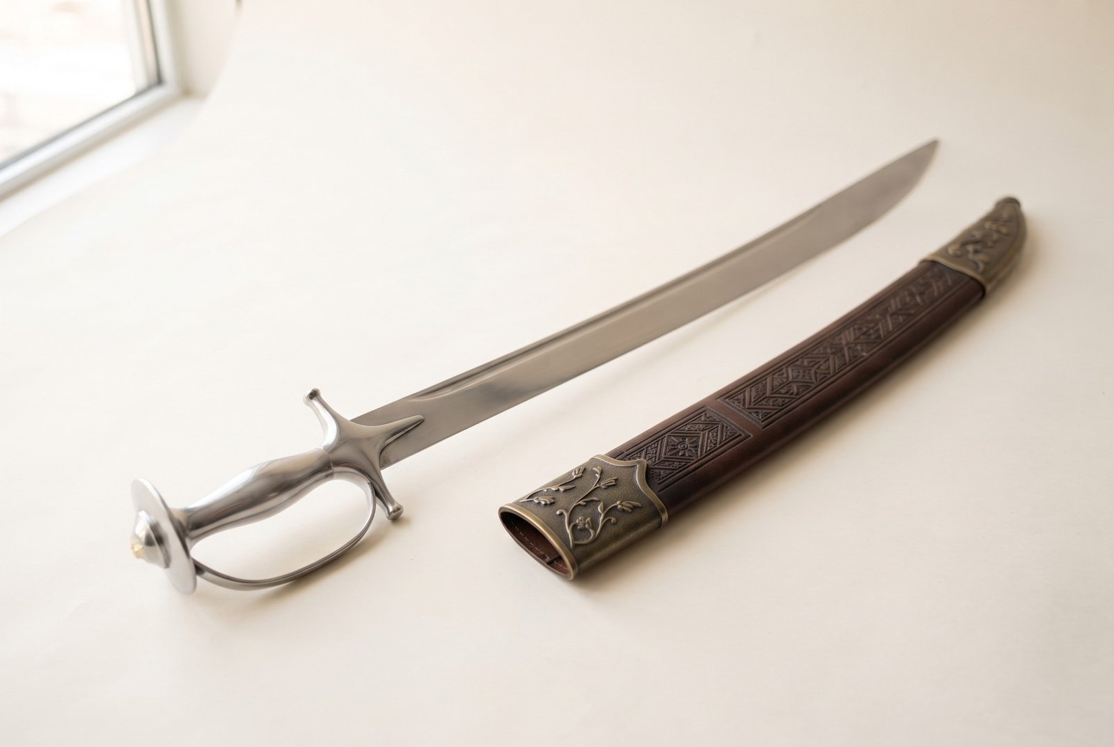 Gemini_Generated_Image_6203rf6203rf6203 Hand forged talwar with a polished curved blade and all steel Indo Persian hilt lying on a pale surface beside a tooled scabbard with chased brass mounts