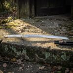 Bone handled wakizashi sword with a bright polished 1095 blade resting on a lichened stone ledge in low sunlight, dark saya lying behind it
