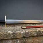 Hand forged Damascus knightly sword with a broad forge welded blade and arched brass cross, resting with its russet scabbard on a lichened sandstone ledge.