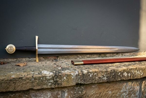 Hand forged Damascus knightly sword with a broad forge welded blade and arched brass cross, resting with its russet scabbard on a lichened sandstone ledge.