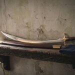 Hand forged shamshir sword with a narrow curved polished blade and one piece rosewood grip resting on a carved timber shelf beside its brass mounted leather scabbard