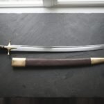 Hand forged Indian sword of talwar type with a mirror polished curved blade and brass disc pommel, laid on dark slate beside its brass mounted leather scabbard